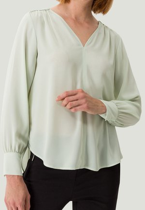 Femme portant un chemisier vert clair à manches longues avec col en V et poignets boutonnés, associée à un pantalon noir, debout devant un fond uni.