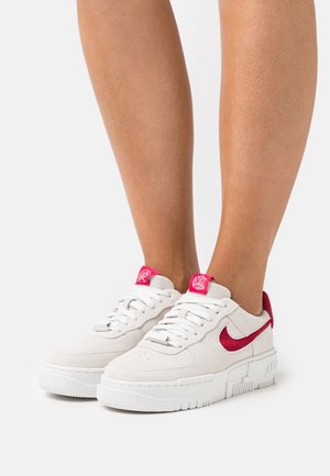 Sneakers in suede bianco con swoosh e dettagli rossi, dotati di una spessa suola bianca e punta testurizzata. I lacci sono bianchi, con un'etichetta del logo sulla linguetta.