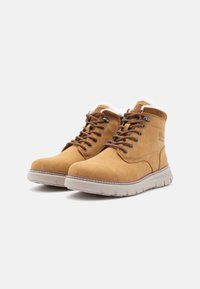 Bocanci de gleznă din suede bej, cu design cu șireturi, guler căptușit, căptușeală din blană sintetică și talpă din cauciuc texturată. Cusăturile în contrast sunt vizibile.