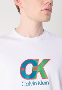 White cotton t-shirt featuring a large multicolored "OK" graphic in blue, green, and red, with "Calvin Klein" printed below in blue.