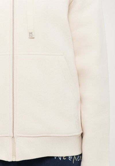 Beige zip-up hoodie with front pocket, drawstring with "WKND" tag, and subtle embroidered logo on lower right, worn over navy pants.