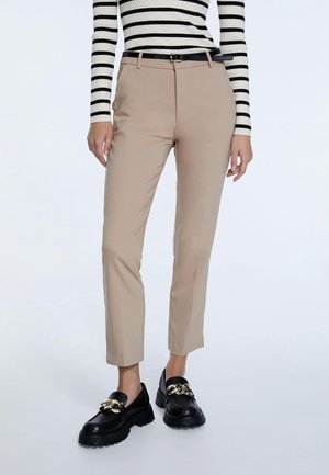 Vrouw die beige slim-fit broek draagt met zwarte riem, zwarte loafers met gouden kettingdetail en een wit-zwart gestreept longsleeve top.