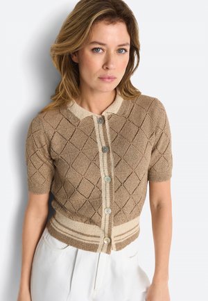 Frau mit blonden Haaren, die eine beige Strickjacke mit kurzem Ärmel und Diamantmuster sowie weiße Hosen trägt und vor einem weißen Hintergrund steht.