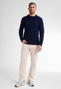 Marineblauer Strickpullover mit geripptem Saum, kombiniert mit hellbeigen Hosen und grauen Turnschuhen, stehend vor einem neutralen Hintergrund.