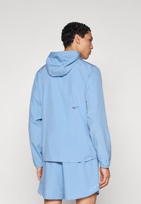 Ανοιχτό μπλε μπουφάν Nike με κουκούλα, μακριά μανίκια, άνετη εφαρμογή και λεπτομέρειες με λογότυπο στην πλάτη. Λεία υφή υφάσματος.