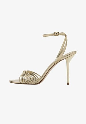 Sandalia dorada de tacón alto con tiras, detalle anudado en la parte delantera, tira delgada en el tobillo y tacón de aguja.