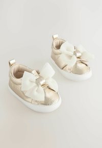 Goldene, metallische Babyschuhe mit großen weißen Schleifen, erhabenem Design und weißen Gummisohlen. Weiche Innenausstattung und Fersenschlaufen für einfaches Anziehen.