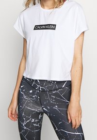 Biały krótki t-shirt z czarnym prostokątnym logo "CALVIN KLEIN". Noszony z czarnymi legginsami z białym marmurowym wzorem i gładką teksturą.