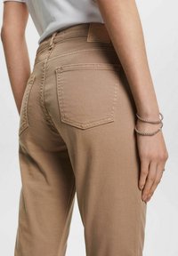 Beige korkeavyötäröiset housut, joissa on muotoon istuva leikkaus, takataskut ja erottuva ompelu. Sujuva pinta, minimaalinen varusteiden käyttö.