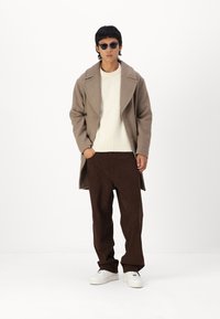 Abrigo de lana marrón, suéter de punto blanco, pantalones de pana marrones y zapatillas blancas. Las gafas de sol añaden un acento oscuro. Silueta simple y estructurada.