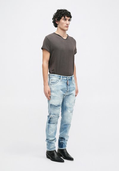 T-shirt v-neck gris foncé, jean skinny bleu clair effet usé avec bord effiloché, patchs usés, et bottines noires. Pose debout contre un fond blanc.