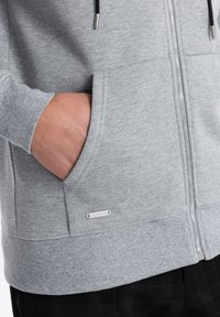 Haut à capuche zippé gris avec des poignets et une taille côtelés, des poches kangourou et un petit logo métallique près de l'ourlet.