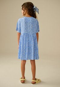 Blauwe bloemenjurk met korte mouwen, een gevormde taille en een vloeiende rok, gecombineerd met gele sandalen met een achterriempje en een gevoerd voetbed.