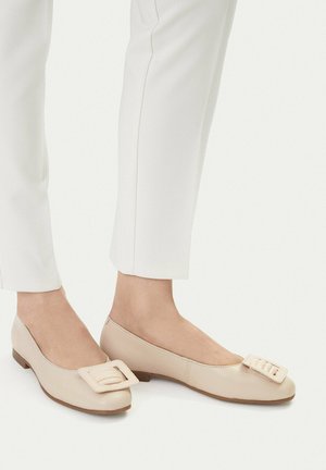 Füße, die beige flache Lederschuhe mit großen quadratischen Schnallen tragen, kombiniert mit weißen Slim-Fit-Hosen vor einem einfarbigen hellen Hintergrund.