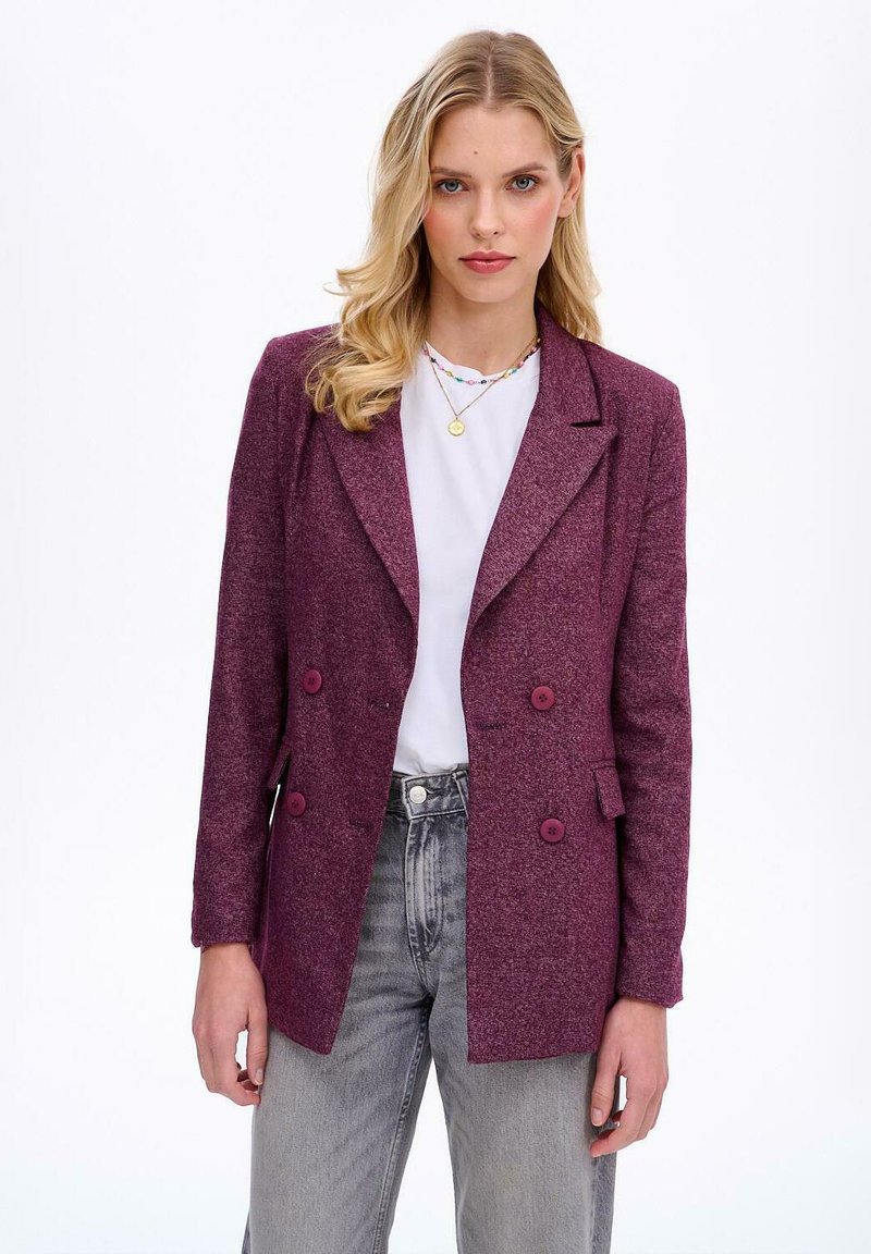 Dwurzędowa marynarka w głębokim purpurowym kolorze z teksturowanym wykończeniem, z klapami oraz dwoma dużymi guzikami, zestawiona z jasnoszarymi dżinsami.