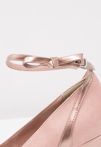 Gros plan sur un escarpin rose à talon haut avec une bride de cheville métallique rose nouée en un petit nœud décoratif.