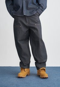 Ciemne dżinsy o szerokich nogawkach, zaakcentowane złotym przeszyciem i przednimi kieszeniami, zestawione z brązowymi butami roboczymi na sznurowadła.