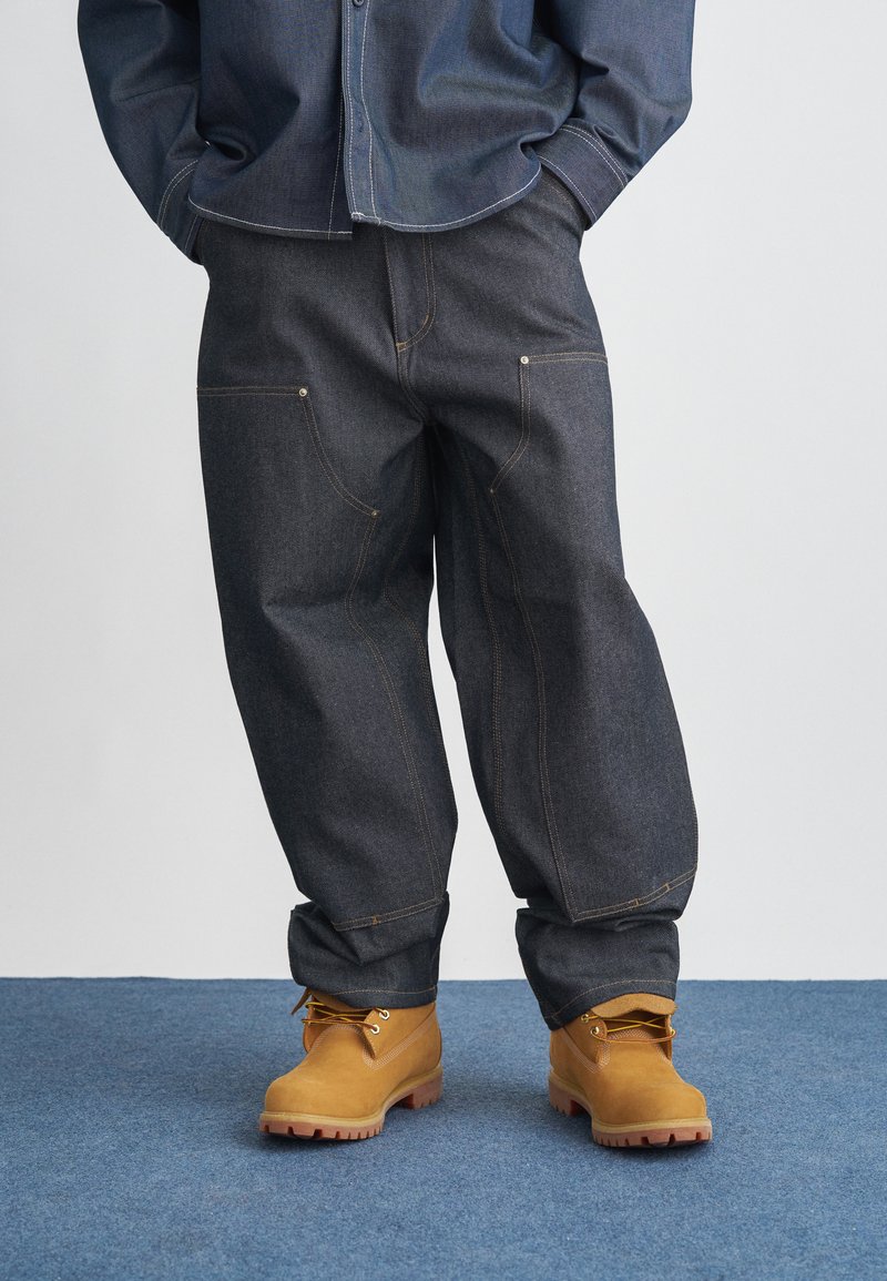Ciemne dżinsy o szerokich nogawkach, zaakcentowane złotym przeszyciem i przednimi kieszeniami, zestawione z brązowymi butami roboczymi na sznurowadła.