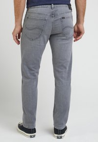 Man wearing gray straight-leg jeans and black Converse sneakers, standing against a plain white background, rear view.