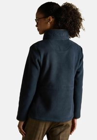 Žena s kučeravými vlasmi, nosiaca okuliare, tmavomodrú flisovú bundu s vysokým golierom a hnedé kockované nohavice, zobrazená zozadu.