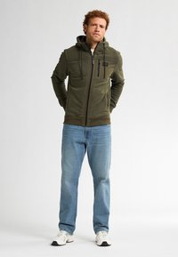 Olijfgroene hoodie met rits en verstelbare capuchon, voorrits en borstzak. Gecombineerd met lichtblauwe jeans en witte sneakers.
