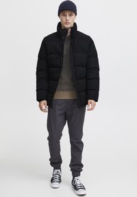 Black padded jacket with high collar, gray sweater, gray pants, and black sneakers. The jacket features a zip closure and a smooth texture.