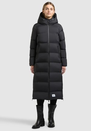 Vrouw staand, draagt een lange zwarte gewatteerde winterjas met capuchon, zwarte legging en zwarte kuitlaarzen op een eenvoudige lichte achtergrond.