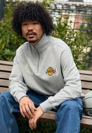 Sudadera de forro polar gris con cuello alto, que presenta un logotipo bordado de "Los Angeles Lakers" en púrpura y amarillo, combinada con jeans azules.