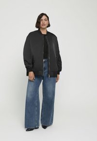Chaqueta bomber negra con cierre de cremallera, bolsillos laterales y puños acanalados, combinada con jeans azules de pierna ancha y zapatos negros de punta.