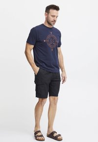 Marine t-skjorte med oransje kompassdesign, kombinert med svarte cargo-shorts. Brune sandaler fullfører det uformelle antrekket.