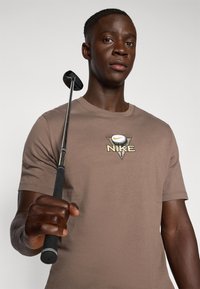 T-shirt en coton marron avec logo Nike, mettant en avant un design de balle de golf. Le modèle tient un club de golf, mettant en valeur une texture douce et une coupe décontractée.
