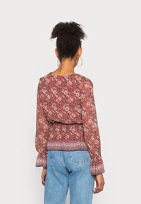 Blusa floreale in ruggine con maniche lunghe a sbuffo, tessuto strutturato e orlo con motivo; abbinata a jeans azzurri chiari.