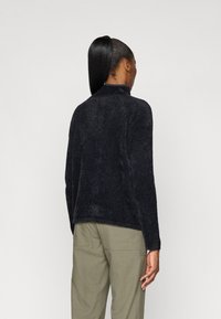 Sort, tekstureret sweater med høj krave og lange ærmer, der har en afslappet pasform og en let buet kant. Sammen med olivenfarvede bukser.