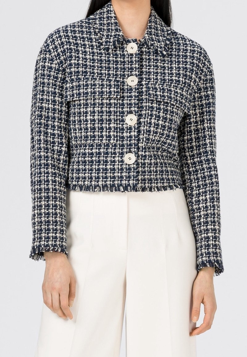 Femme portant une veste courte en tweed à carreaux bleu marine et blanc avec de gros boutons blancs et un pantalon blanc taille haute.