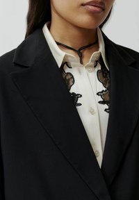 Zwarte blazer met een gestructureerd ontwerp, gecombineerd met een witte blouse met zwarte kantdetails op de kraag en een zwarte koord choker.