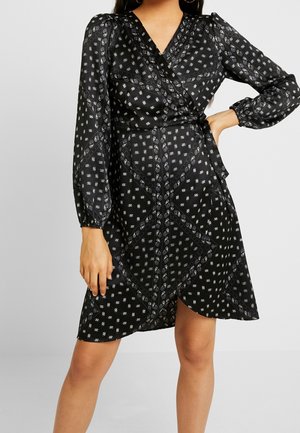 Femme portant une robe portefeuille noire avec des motifs paisley et floraux blancs, des manches longues bouffantes et une taille nouée, debout avec une main sur la hanche.