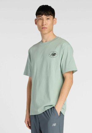 Camiseta de algodón verde menta con mangas cortas, cuello redondo y un logo negro en el pecho. Combinada con pantalones cortos deportivos grises.