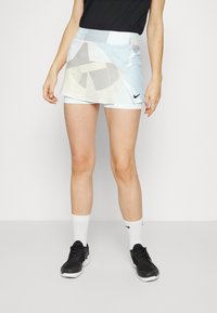 Falda deportiva para mujer de Nike, de color azul claro con acentos de patrón geométrico. Presenta una cintura alta y shorts internos. Zapatillas negras y calcetines blancos.