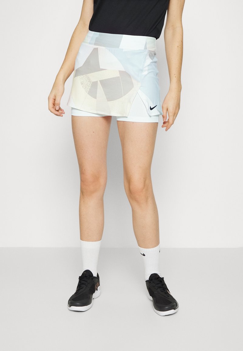 Falda deportiva para mujer de Nike, de color azul claro con acentos de patrón geométrico. Presenta una cintura alta y shorts internos. Zapatillas negras y calcetines blancos.