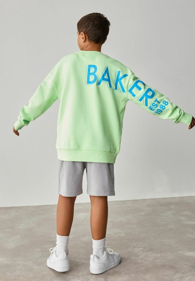 Mintgroen sweatshirt met blauwe tekst "BAKER" op de achterkant, grijze shorts, witte sneakers en witte sportsokken. Glad stof, relaxte pasvorm.
