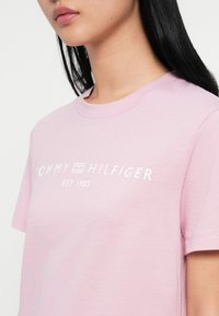 Ružová bavlnená tričko s okrúhlym výstrihom, krátkymi rukávmi a bielym textom "TOMMY HILFIGER EST 1985" napísaným cez hruď.