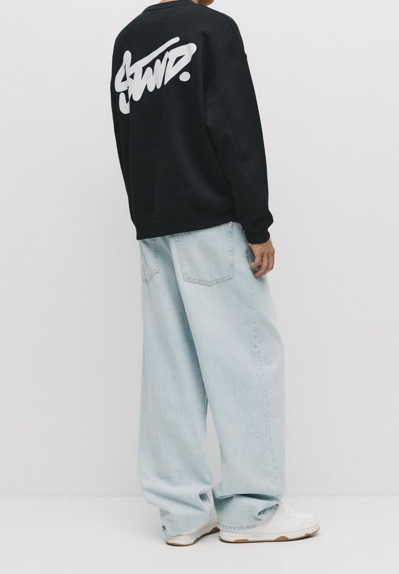 Personne portant un sweat noir avec un texte blanc stylisé dans le dos, un jean bleu clair à jambes larges et des baskets blanches, debout devant un fond uni.