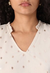 Blusa beige chiara con piccole macchie metalliche dorate, colletto arricciato dettagliato e design a V, realizzata in un morbido tessuto testurizzato.