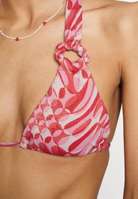 Top de biquíni com formato triangular, padrões abstratos em rosa e vermelho, textura cintilante e detalhe em anel circular na alça do pescoço.