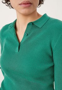 Grün geripptes Polohemd mit Kragen, zwei Knöpfen am Hals und strukturiertem Strickstoff. Nahaufnahme, die das Design des Hemdes hervorhebt.