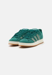 Grüne Wildledersneaker mit niedrigem Profil, ausgestattet mit tonalen Schnürsenkeln, drei seitlichen Streifen und einer crèmefarbigen Gummisohle. Akzentuiert durch ein Logo auf der Zunge.