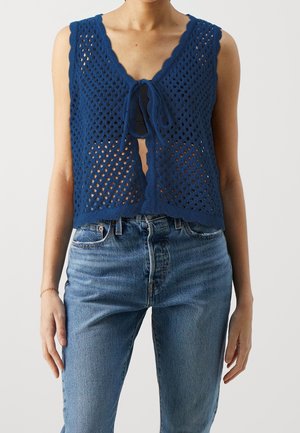 Veste sans manches en maille ajourée bleue nouée à l'avant sur un haut noir, associée à un jean taille haute bleu clair, portée par une personne debout.