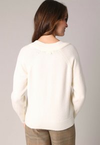 Femme aux longs cheveux bruns portant un pull en tricot crème et un pantalon à carreaux marron, vue de dos contre un fond uni.