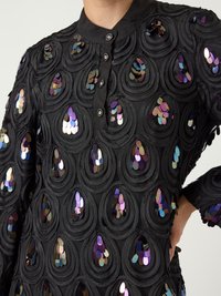 Μαύρο ένδυμα με υφασμάτινη υφή και κυκλικά σχέδια, διακοσμημένο με ιριδίζουσες παγιέτες, με κουμπί στο γιακά.