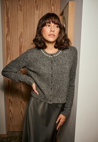 Cárdigan de lana gris con cuello redondo, adornado con pequeños elementos decorativos, cierre de botones y puños acanalados, llevado sobre una falda de satén oscura.
